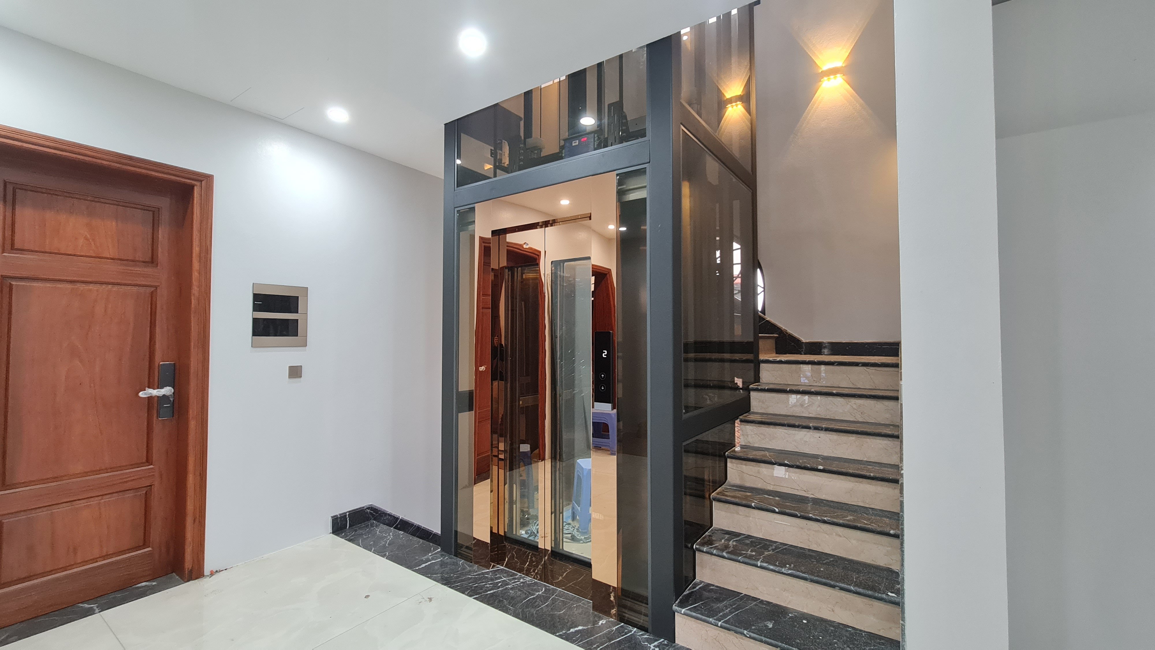 Thang máy gia đình giếng vách kính trà khói, cửa cabin đồng cạnh cầu thang đá, biệt thự Văn Giang