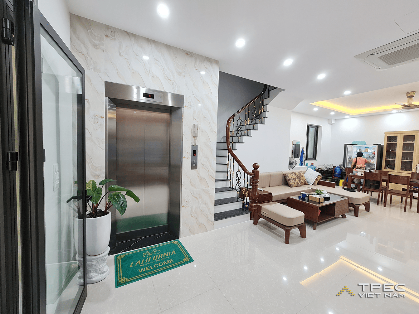 Thang máy gia đình cửa inox đặt cạnh cầu thang gỗ trong phòng khách nhà phố 5 tầng ở Hoàng Mai