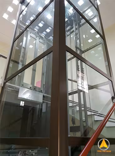 Giếng thang máy vách kính khung nhôm màu nâu đồng cạnh cầu thang, nhìn từ dưới lên