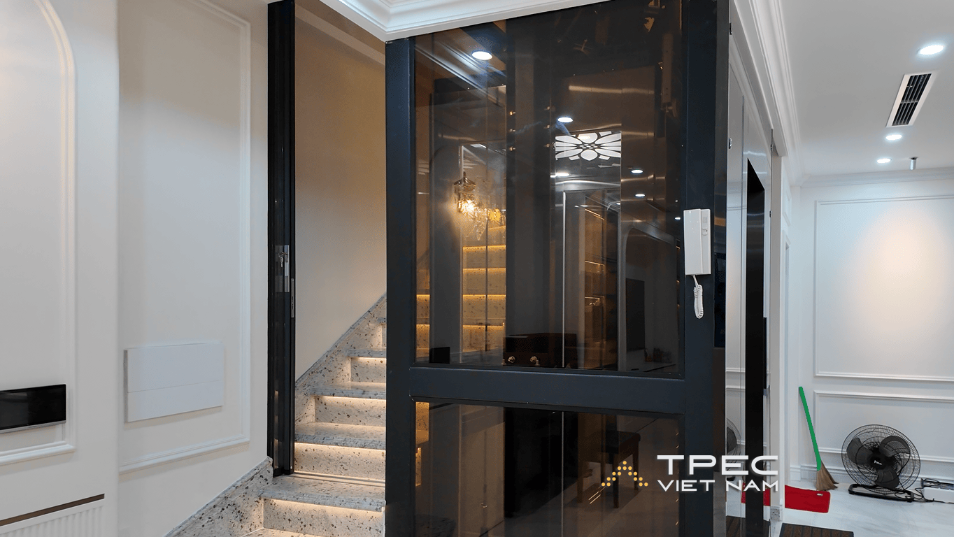 Thang máy kính khung inox gương đen đặt cạnh thang bộ ốp đá trong nhà phố tại Trung Kính