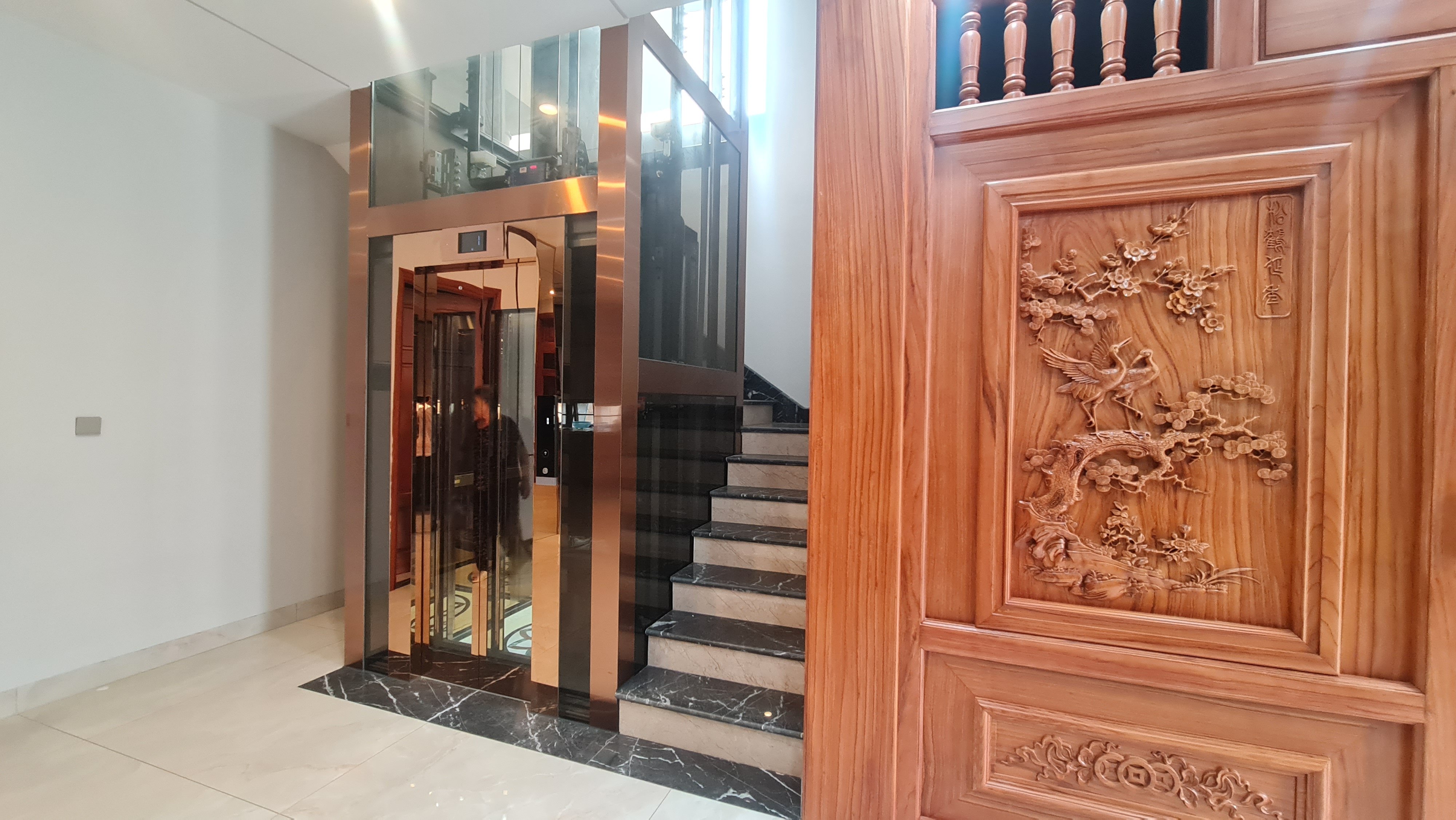 Thang máy gia đình khung inox màu đồng cạnh cầu thang đá và tranh gỗ chạm, biệt thự Văn Giang