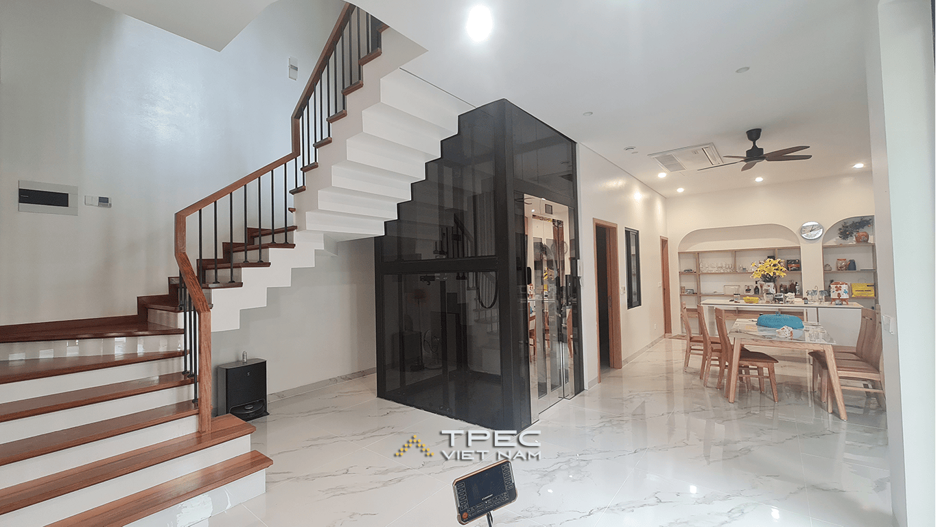 Không gian tầng 1 nhà phố Đông Anh với thang máy kính khung đen đặt cạnh cầu thang, liền phòng bếp