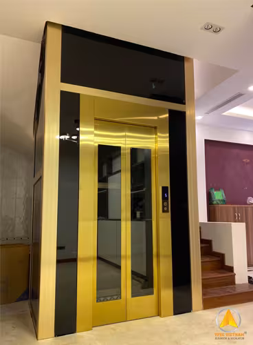Cabin thang máy cửa inox mạ vàng gương, giếng khung nhôm kính đen sang trọng đặt cạnh cầu thang
