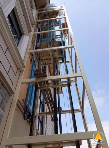 Khung thép thang máy sơn màu kem lắp ngoài trời sát tường bên tòa nhà, nhìn từ dưới lên