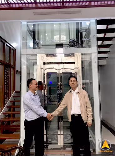Bàn giao thang máy gia đình khung thép vách kính lắp cạnh cầu thang trong nhà