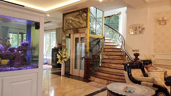 Thang máy gia đình cabin kính trong sảnh biệt thự có cầu thang xoắn và bể cá cảnh